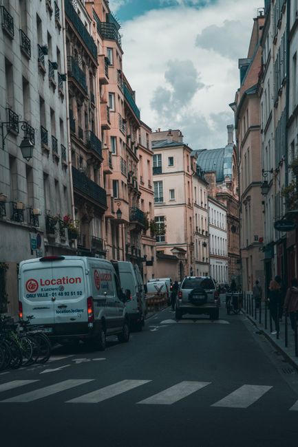 A red van belonging to Man and Van Edgware is parked on a cobblestone street in front of a multi-story residential building with a pinkish-brown facade, large windows, and decorative architectural details, including a sculpture mounted on the exterior wall. The van is positioned close to the building's entrance and partially blocks a 'no entry' traffic sign, with a bicycle leaning against a nearby wall and a wooden bench situated on the pavement. The surrounding environment includes a street lamp, bollards, and a narrow alley visible behind the van, indicating an urban setting suitable for house removals, furniture transport, and home relocation activities, especially in narrow streets and apartment complexes facilitated by professional moving services.