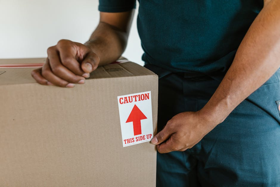 Close-up view of a person wearing a dark green t-shirt and dark trousers, holding a cardboard box during a home relocation process. The individual's hands are gripping the box, which is sealed with pa