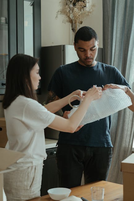 Close-up view of a person wearing a dark green t-shirt and dark trousers, holding a cardboard box during a home relocation process. The individual's hands are gripping the box, which is sealed with pa